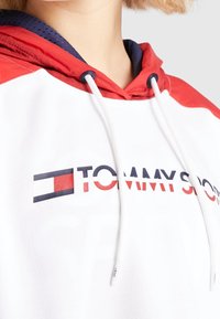 Primo piano di una persona che indossa una felpa Tommy Sport bianca e rossa con lacci bianchi e una fodera interna blu.