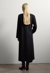 Cappotto lungo nero con un design strutturato, taglio dritto e maniche oversize. Il materiale appare liscio, elegante e scende oltre le ginocchia.