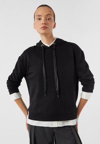 Czarna bluza z kapturem i sznurkami, z długimi rękawami i luźnym krojem. Założona na białą koszulę z kołnierzykiem, miękka tekstura, minimalistyczny design.