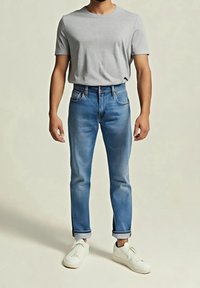 Homme portant un t-shirt gris clair à manches courtes, un jean bleu retroussé et des baskets blanches, debout devant un fond clair uni.