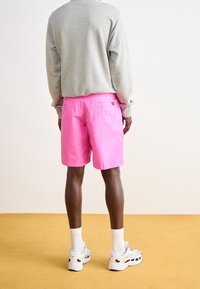Pink cotton shorts with a relaxed fit, functional back pockets, and a tag detail, paired with a gray sweatshirt and white athletic shoes.