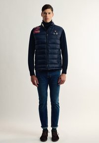 Chaleco acolchado azul marino con cierre de cremallera, diseño de acolchado horizontal, parches bordados, llevado sobre un suéter de cuello alto negro, combinado con jeans azules y zapatos marrones.