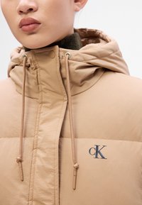 Béžová péřová bunda s kapucí, která má hladký povrch a mírný lesk. Obsahuje stahovací šňůrky a tmavě modré logo "cK" na hrudi.