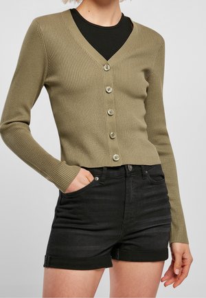 Kvinde iført en olivengrøn ribbet cardigan med knapper over en sort top, kombineret med sorte denimshorts med oprullede kanter, med den ene hånd i lommen.