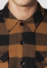 Camicia a quadri marrone e nera con collo, chiusura con bottoni e taschino sul petto in tessuto intrecciato e strutturato.