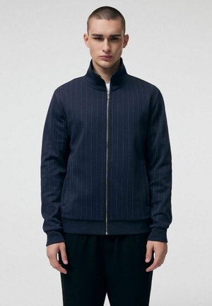 Młody mężczyzna o krótkich włosach, ubrany w granatową kurtkę na zamek z subtelnymi pionowymi prążkami, stojący na tle jasnego, jednolitego tła.