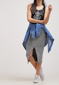 Débardeur noir avec broderie blanche, jupe midi grise avec une fente devant, et veste en denim nouée autour de la taille, associée à des baskets blanches.