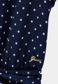 Meri sininen kangas valkoisilla pilkuilla, jossa on kerrostettu yksityiskohta ja kultainen "Guess"-logo helman kohdalla. Sileä pinta.