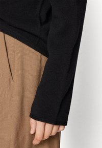 Schwarzer Strickpullover mit geripptem Muster an den Ärmeln, kombiniert mit hellbraunen Hosen, die Falten und Taschen haben. Einfaches, lässiges Design.