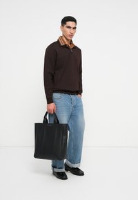 Άνδρας φορέσει καφέ πουλόβερ, ανοιχτό μπλε τζιν και μαύρα παπούτσια, κρατά μια μαύρη τσάντα tote με χερούλια, ενώ στέκεται σε λευκό φόντο.