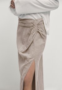 Jupe en lin beige avec une fente devant et un détail de nœud torsadé à la taille. Associée à un haut en tricot texturé de couleur claire.