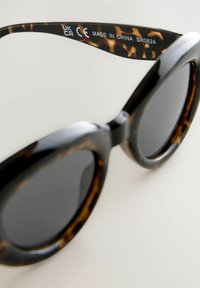 Gafas de sol redondas con montura negra que presenta un patrón de carey, lentes oscuros y texto grabado en la patilla.