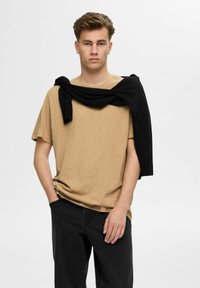 Beige short-sleeve t-shirt with a relaxed fit, paired with black sweater draped over shoulders, and dark jeans. Smooth texture, casual design.