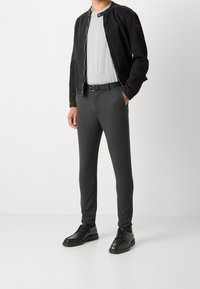 Man in een zwarte ritsjack, lichtgrijs t-shirt, donkergrijze slim-fit broek, zwarte riem en zwarte veterschoenen, staand met handen in de zakken.