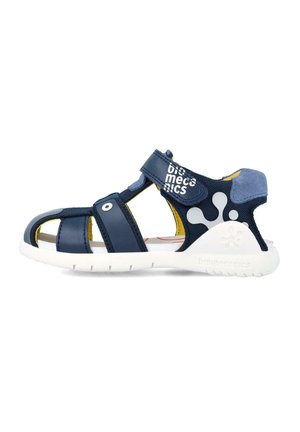 Sandalia azul marino con material de malla y cuero, diseño abierto, cierre de strap con Velcro, acentos amarillos y suela de goma blanca con detalle de marca.