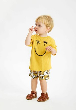 Gul bomulls T-shirt med en svart smiley och palmträd design, ihop med vita shorts med ett bananmönster, bruna sandaler.