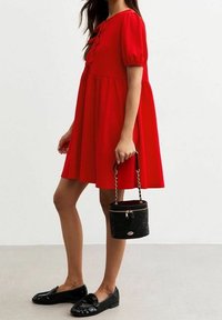 Robe rouge à manches bouffantes avec une taille cintrée, tissu texturé et détails de boutons. Associée à des mocassins noirs en vernis et un sac matelassé rond.
