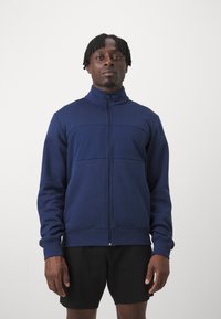 Marinen blå lynlåsjakke lavet af blødt materiale, med høj krave, brystpanel og ribbede manchetter, parret med sorte shorts.