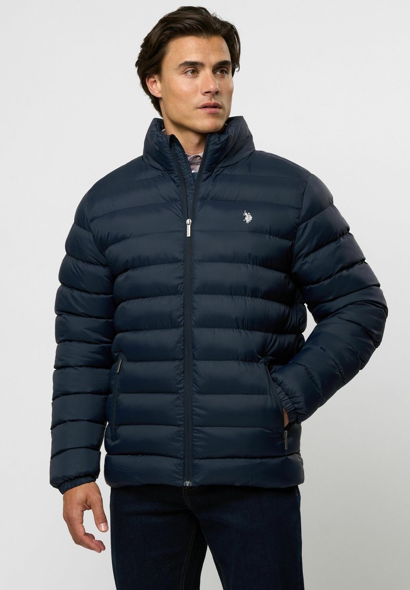 Noor mees, kes kannab tumesinist pusa kõrge kraega ja tõmblukuga taskutega, seisab ühevärvilise hele tausta ees.