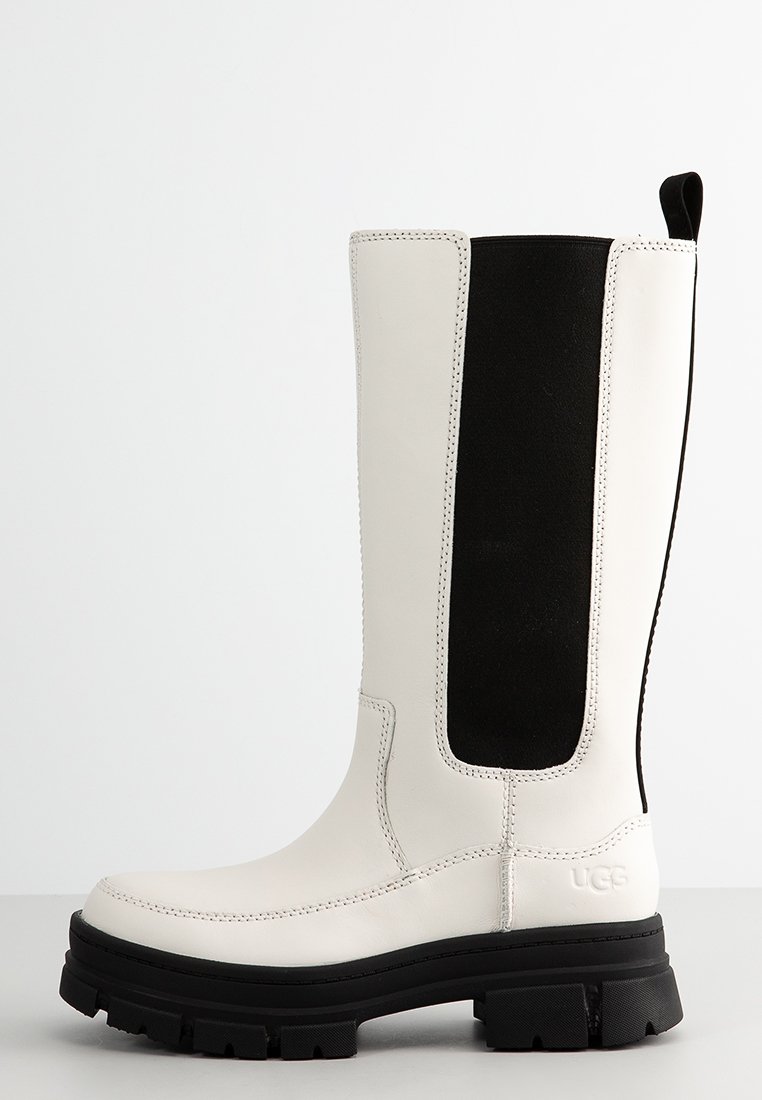 Weiße lederne Stiefel bis zum Knie mit einem schwarzen elastischen Seitenpanel, strukturiertem Sohlen und kontrastierender Naht. Mit UGG-Branding an der Ferse.