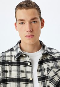 Geruite flanellen shirt in zwart en wit, met knopen, een kraag en een borstzak. Draagt over een wit T-shirt met ronde hals.