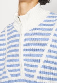 Primo piano di un maglione a costine bianco con righe blu, collo alto e cerniera argentata parzialmente chiusa.