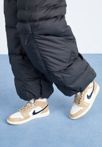 Osoba v béžových a bílých teniskách Nike a černých nafouklých kalhotách stojí na světle modré kobercové podlaze.