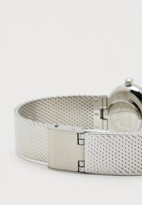 Zilveren metalen horlogeband van gaas met een veilige sluiting, voorzien van een gegraveerd rechthoekig ontwerp, opgerold en plat gelegd op een witte achtergrond.