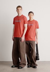 Dos personas llevan camisetas de manga corta color coral con el logo "tommy" a juego. Combinan con pantalones marrones holgados y zapatos negros, y llevan un bolso.