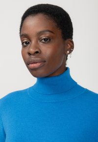 Maglione blu a collo alto in morbido tessuto a coste. Il modello indossa più orecchini. Lo sfondo è bianco uniforme.