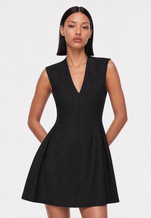 Frau trägt ein ärmelloses schwarzes V-Ausschnitt-Kleid mit tailliertem Oberteil und ausgestelltem Rock, steht vor einem einfarbigen hellen Hintergrund.