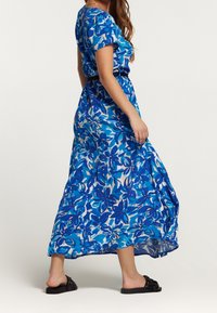 Donna che indossa un abito midi floreale blu con una cintura nera, che cammina scalza in sandali neri, su uno sfondo bianco uniforme.