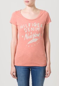 Camiseta de algodón color durazno con mangas cortas, cuello redondo y texto impreso en blanco "HILFIGER DENIM New York" con un gráfico de pluma.