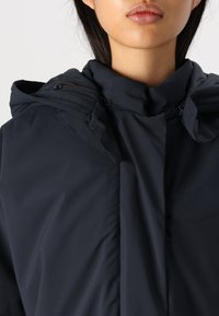 Veste isolante bleu marine foncé avec un col montant, des accents de boutons et une capuche amovible avec des détails plissés. Texture de tissu lisse.