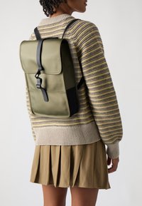 Sac à dos vert olive avec un rabat repliable, des bretelles noires et une fermeture à bouclerie. Matériau lisse, forme rectangulaire et design minimaliste.
