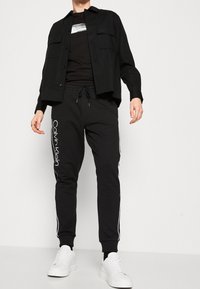 Zwarte button-up shirt, zwarte t-shirt met logo, zwarte sweatpants met witte strepen en witte sneakers. Casual outfit met sportieve details.