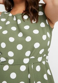 Grünes Kleid mit großen weißen Polka-Dots, ärmelloses Design, strukturierter Stoff, tailliert mit einem Gürtel und dezenten Falten.