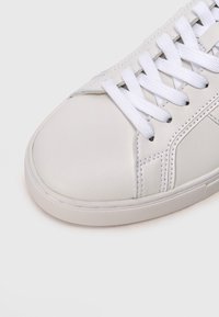 Κοντινό πλάνο του μπροστινού μέρους ενός λευκού αθλητικού παπουτσιού με λευκά κορδόνια, εμφανή ραφές και υφή στη σόλα, σε απ�λό φόντο.