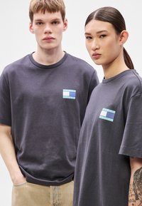 T-shirts de algodão cinzentos apresentam um pequeno emblema azul e branco no peito. O tecido parece macio, com um ajuste descontraído e mangas curtas.