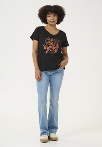 T-shirt noir avec un col en V et un motif floral cuivré. Associé à un jean évasé bleu clair et des chaussures beiges. Texture plate et lisse.