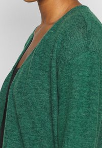 Zielony, dziergany kardigan o luźnym kroju, z dekoltem w serek i prążkowaną fakturą, zaprezentowany pod kątem bocznym.