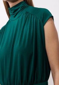 Kvinna i en ärmlös, rynkad grön topp med hög hals och lös passform mot en ljus bakgrund.