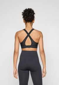 Black sports bra with crisscross straps, textured fabric, and Under Armour logo along the bottom. Paired with high-waisted black leggings.