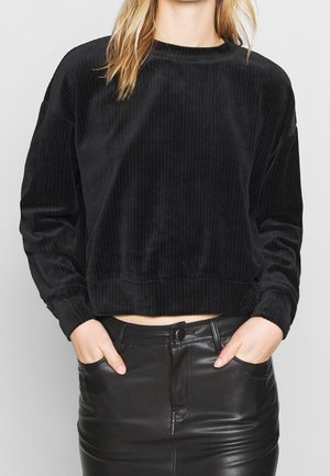 Vrouw draagt een zwarte geribbelde fluwelen trui met lange mouwen en een zwarte leren rok, met handen in de zakken, afbeelding bijgesneden bij nek en heupen.