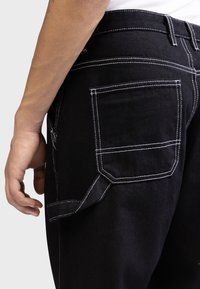 Pantaloni in denim nero con cuciture bianche, dotati di tasca posteriore e dettaglio a cappio laterale. Texture liscia; vestibilità slim in vita.