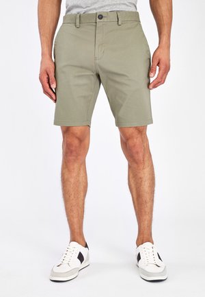Shorts de color verde oliva para hombre, hechos de una mezcla de algodón, con frente plano, lazos para cinturón y cierre de botón. Combinados con zapatillas blancas.