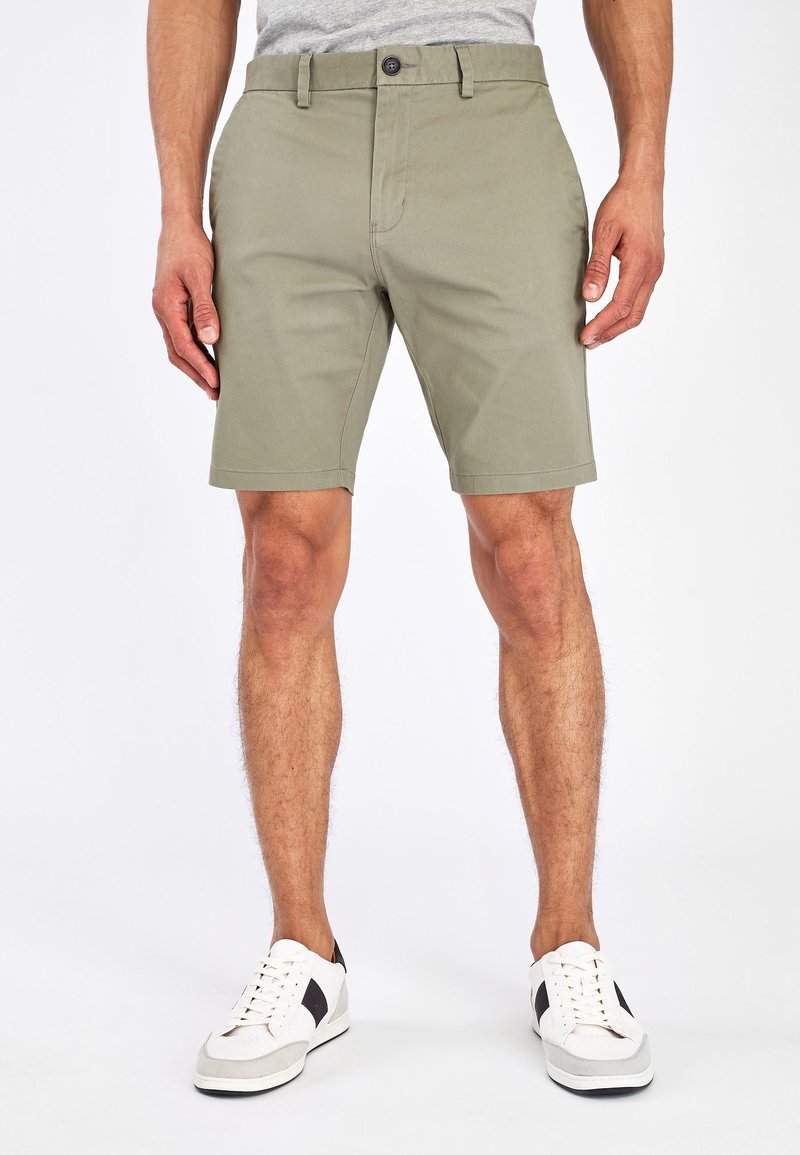 Shorts de color verde oliva para hombre, hechos de una mezcla de algodón, con frente plano, lazos para cinturón y cierre de botón. Combinados con zapatillas blancas.