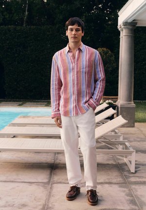 Young man stands by poolside wearing a multicolored striped shirt, white pants, and brown shoes, with lounge chairs and greenery in the background.