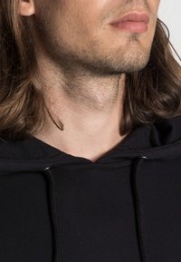 Felpa nera con cappuccio e cordoncino, caratterizzata da una morbida trama del tessuto. In primo piano si vedono la zona del collo e i capelli, mettendo in evidenza i dettagli del collo del capo.