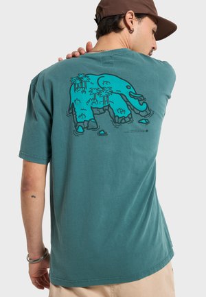T-shirt di cotone color teal con grafica di un elefante blu sul retro, circondato da palme tropicali, con vestibilità comoda e maniche corte.